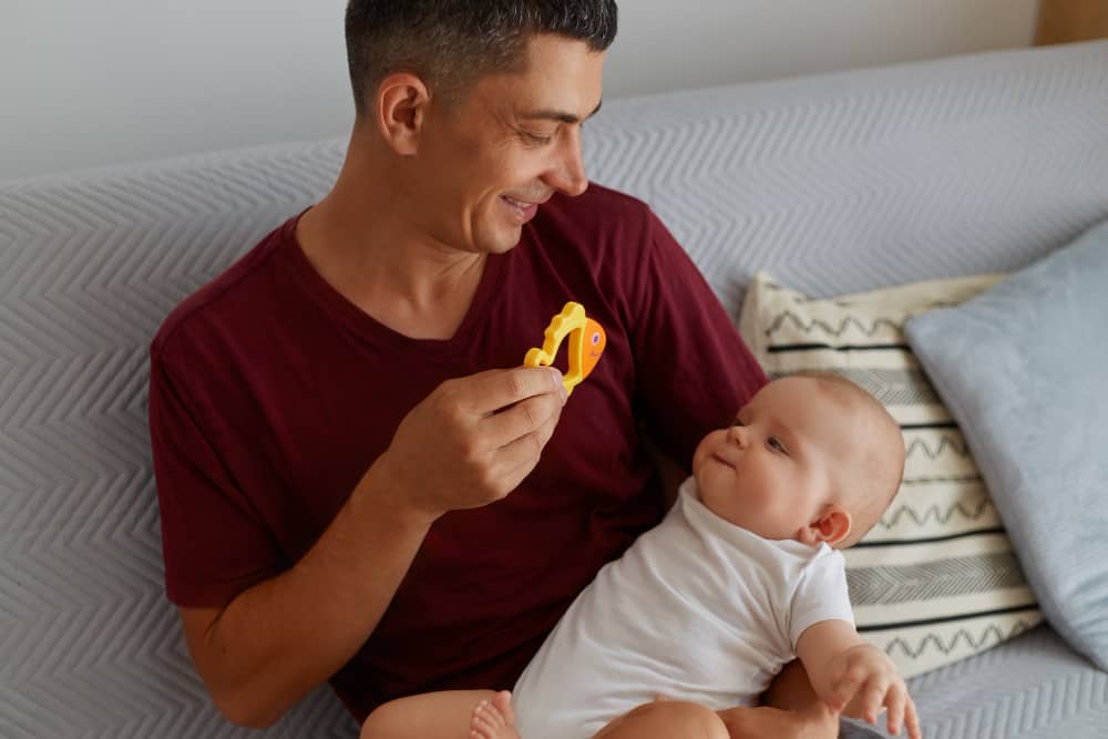 happy father holding toy playing with his baby boy girl while sitting sofa smiling man wearing maroon t shirt showing child orange fish happy parenthood min - paternity leave in UK in General